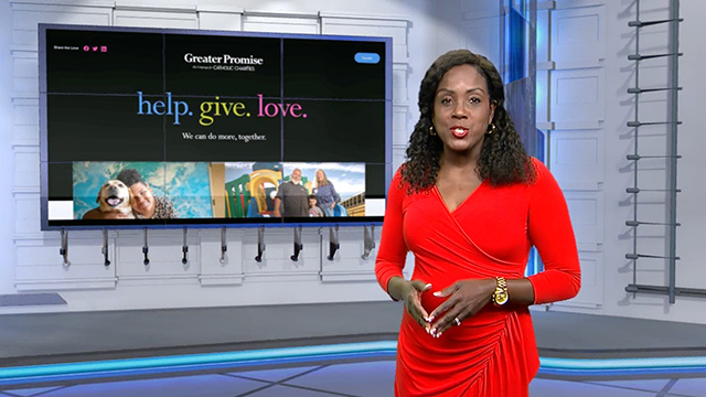 News anchor in a red shirt standing in front of a screenshot of the Greater Promise website