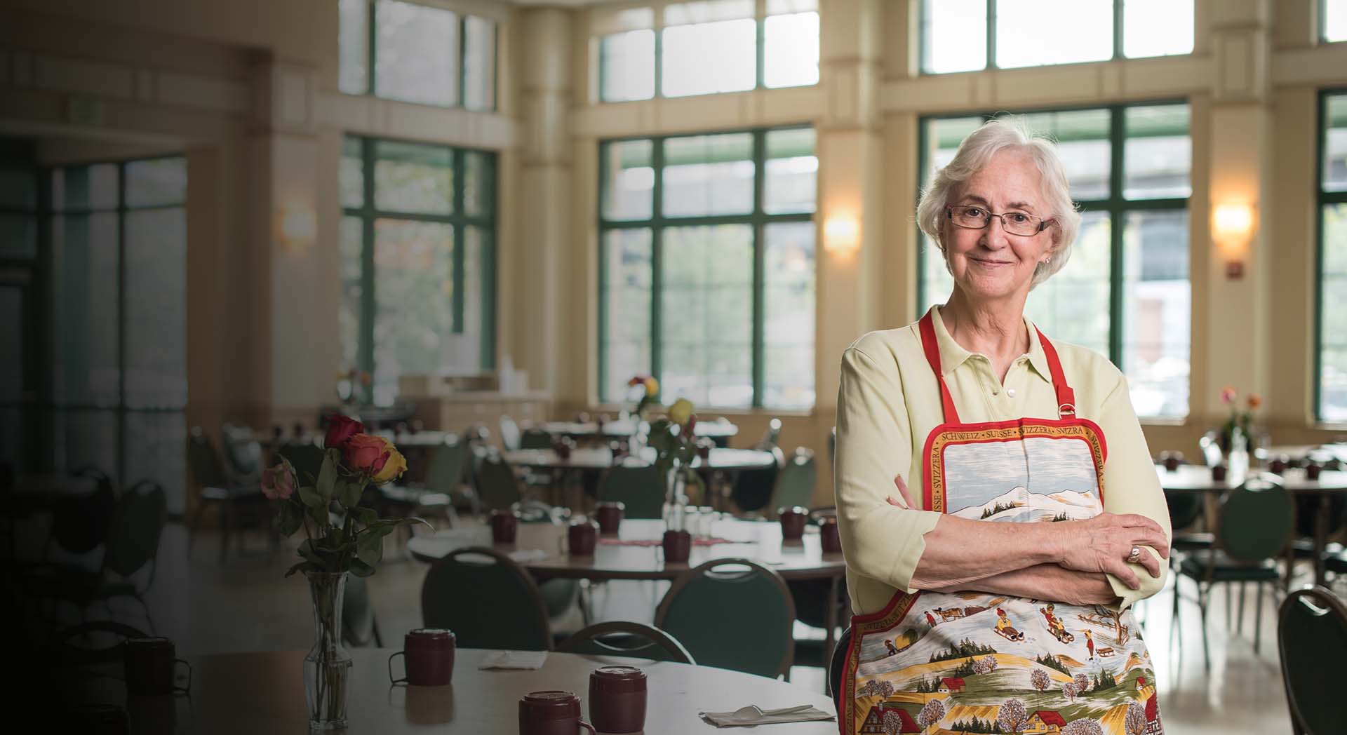 older lady in empty cafeteria wearing an apron with arms folded