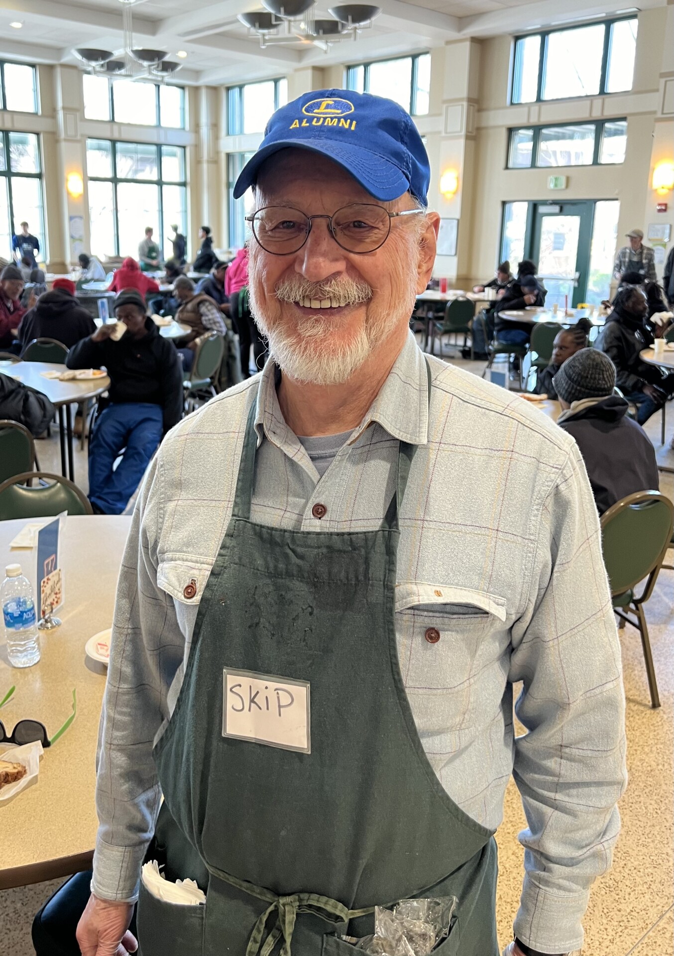A man wearing an apron and a baseball cap smiles in a cafeteria.
