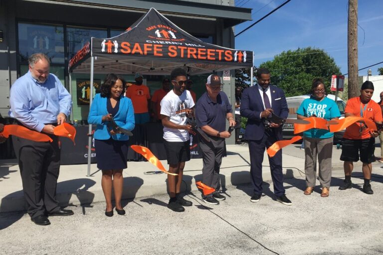 Community leaders stand in front of the new Safe Streets building with large scissors while cutting a large orange ribbon