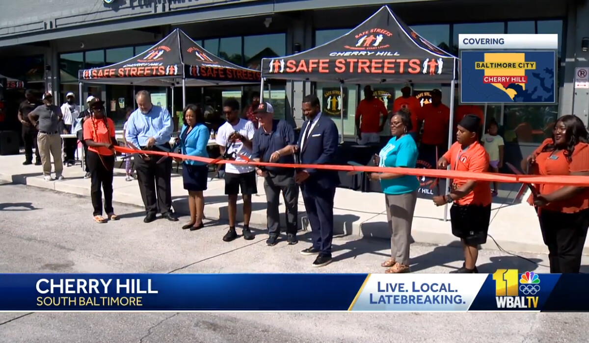 Baltimore community leaders stand beside each other in front a new office building with large scissors as they cut a large orange ribbon